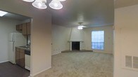 This single-floor residence offers a comfortable and functional layout designed for everyday living. The main living area is a generous, open space finished with plush wall-to-wall carpeting that adds warmth underfoot. A standout architectural feature is the corner fireplace, accented by a raised brick hearth and a clean white surround with a distinct angled chimney hood that draws the eye upward, serving as a natural focal point in the room. The textured ceiling adds subtle character throughout the space, while ceiling fans provide efficient air circulation and overhead lighting. A window positioned near the fireplace allows natural light to filter into the living area, brightening the neutral-toned walls and contributing to an open, airy feel. The light beige color palette across the walls, ceiling, and carpet creates a cohesive, understated aesthetic that lends itself well to a variety of interior styles. Overall, this space presents a straightforward, well-proportioned single-level layout with practical features and a warm, inviting ambiance suitable for comfortable residential living.