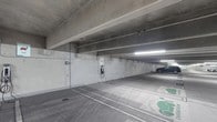 This single-level covered parking facility is constructed with a robust exposed concrete structure, featuring solid load-bearing columns and a flat overhead deck that provides full weather protection. The space is designed with practicality and modern functionality in mind, offering a clean, well-organized layout with clearly marked parking bays and directional lane markings painted directly on the smooth concrete floor. A notable feature of this facility is the inclusion of dedicated electric vehicle (EV) charging stations, mounted directly to the concrete walls and clearly identified with EV-designated floor markings, reflecting a forward-thinking approach to sustainable transportation infrastructure. Overhead fluorescent strip lighting runs the length of the facility, ensuring consistent and reliable illumination throughout the interior even in the absence of natural light. The exposed concrete ceiling reveals structural beams and utility conduits, giving the space an industrial yet functional aesthetic. The facility is designed to accommodate multiple vehicles with an open, column-supported layout that allows for straightforward navigation and parking. Overall, this parking facility combines structural durability with modern amenities, making it a practical and well-equipped space for everyday use.