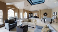 This single-floor space makes an immediate impression with its dramatic vaulted ceilings and a bold, teal-accented skylight that serves as a stunning architectural centerpiece. The open-concept layout is bathed in natural light, courtesy of a series of tall arched windows that line the walls and invite views of the surrounding greenery outside. Light-toned tile flooring extends throughout, enhancing the sense of openness and continuity across the space. The main lounge area is furnished with a comfortable arrangement of cream sofas and upholstered accent chairs, anchored by a sleek dark coffee table — creating a welcoming environment for gathering and relaxation. Additional seating and dining areas are thoughtfully arranged throughout, offering flexible zones for various uses. Neutral warm-beige walls provide a grounding backdrop that complements the variety of furnishings. French-style glass doors connect the interior to adjacent areas, adding both elegance and functionality to the flow of the space. Recessed lighting supplements the natural illumination, ensuring the space remains bright and inviting at all hours. Overall, the space blends classic architectural details with contemporary furnishings to create a refined, comfortable, and highly functional environment.