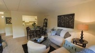 This single-floor residence is designed with an open-concept layout that seamlessly connects the living, dining, and kitchen areas into one cohesive space. The living area is anchored by a plush, cream-colored sofa and loveseat arrangement, complemented by a dark, patterned ottoman that doubles as a coffee table. Decorative throw pillows in muted teal and brown tones add visual interest, while a set of four dark-framed botanical canvas prints adorns the wall, providing an elegant focal point. A black ladder-style bookshelf adds vertical dimension and storage to the space. The dining area accommodates a round dark-wood table with matching chairs, positioned adjacent to the kitchen pass-through window. The kitchen features white cabinetry, offering a clean and classic aesthetic. Neutral-toned carpeting throughout the living and dining areas contributes to the warm, understated ambiance of the home. Natural light filters into the space, enhancing the light wall tones and creating a welcoming atmosphere. The overall design balances functionality with tasteful décor.