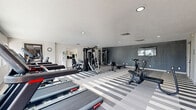 This fitness center offers a generously sized, single-floor workout facility designed to accommodate a full range of exercise needs. The space is finished in a clean, contemporary style, highlighted by a bold gray-and-white striped vinyl tile floor that adds visual energy to the room. Recessed lighting and a textured ceiling complement the bright, neutral-toned walls, creating an open and motivating environment.

The cardio zone is well-stocked, featuring multiple treadmills and an elliptical trainer positioned along a windowed wall that brings in ample natural light and outdoor views — ideal for energizing workouts. Strength training is equally well-represented, with a multi-station cable machine, rowing machines, stationary bikes, and a dedicated free weight and bench area. A wall-mounted television provides entertainment for users during their sessions.

Separate restroom facilities are accessible directly from the main workout floor, adding convenience for users. A secondary door provides additional access and ventilation to the space. The overall layout is thoughtfully organized to allow clear circulation between equipment zones, ensuring a comfortable and functional workout experience. The combination of modern finishes, abundant equipment variety, and practical amenities makes this fitness center a comprehensive facility for both cardio and strength training enthusiasts.