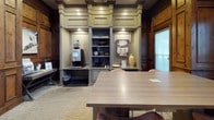 This single-story business center is defined by its striking dark wood panel millwork, which lines the walls from floor to ceiling throughout the space, lending a warm, classic character to an otherwise contemporary work environment. The paneling is detailed with raised moldings and framed sections, creating a sense of architectural depth and craftsmanship that sets the tone for the entire center.

The workspace area features a long, smooth work surface paired with modern ergonomic seating in warm burnt-orange tones, providing comfortable seating for multiple users. An integrated built-in bookshelf along the base of the workstation adds a practical touch. A large window admits natural light, softened by sheer window treatments that maintain privacy while brightening the interior.

A dedicated lounge area offers two upholstered striped armchairs accented with vibrant orange throw pillows, positioned near a large interior window and a solid wood entry door—creating an inviting spot for informal conversation or relaxed work.

A separate area houses a built-in display and refreshment station, complete with a coffee machine, decorative shelving, and accent lighting, rounding out the amenities of this polished and well-appointed business center.