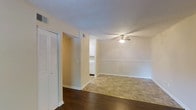 This single-story residence at Iroquois Gardens offers a straightforward and functional layout designed for comfortable everyday living. The main living area is finished with warm beige walls and white trim, creating a clean, neutral palette throughout. A ceiling fan centered in the living space provides both light and air circulation, contributing to the room's practical comfort.

The flooring transitions noticeably between areas, with dark hardwood-style flooring in the foreground giving way to light tile in the main living zone, adding visual interest and helping to define distinct functional areas within the open layout. White louvered bi-fold closet doors along one wall provide generous storage while maintaining a cohesive, classic aesthetic.

A textured ceiling adds subtle depth overhead, and HVAC vents are positioned for efficient climate control throughout the space. The open passageway leading toward the kitchen area is visible in the background, where white cabinetry can be glimpsed, suggesting a clean and functional kitchen. The overall ambiance is bright, neutral, and move-in ready, offering a blank canvas well-suited for personalization.