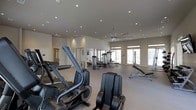 This fitness center offers a thoughtfully designed workout space spread across a single floor. The room features a neutral color palette with light gray walls and a smooth, light-toned flooring that contributes to a clean, modern aesthetic throughout. Recessed lighting is installed across the ceiling, providing consistent, even illumination, and is complemented by a large ceiling fan that promotes air circulation during workouts.

Large windows along the far wall allow natural light to fill the space, creating an open and energizing atmosphere. The layout is spacious, with equipment arranged to allow comfortable movement between stations. Cardio machines, including ellipticals, are positioned prominently toward the front of the room, while a selection of cable and weight machines occupy the middle and rear areas. A padded adjustable bench is also available for strength training exercises.

Full-length mirrors are mounted along one wall, enhancing the sense of space and allowing users to monitor their form. A separate door visible toward the rear may indicate access to an additional room or entry point. Overall, the fitness center is well-organized, functional, and designed to support a broad range of workout routines.
