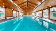 Located in Calgary, Alberta, this indoor pool facility is a well-appointed aquatic space defined by its distinctive architectural character and bright, welcoming atmosphere. The centerpiece is a generously sized lap pool with crystal-clear turquoise water, bordered by wide, light-toned tiled decking that provides ample space for swimmers and spectators alike.

The most striking architectural feature is the soaring vaulted ceiling clad in warm natural wood, supported by elegantly curved wooden arches that frame the space with a sense of grandeur. A row of clerestory windows runs along the roofline, flooding the interior with abundant natural light and enhancing the open, airy feel of the room. Additional pendant light fixtures hang from the ceiling, ensuring the space is well-lit at all times.

Large windows line both sides of the facility, offering views of the surrounding outdoor greenery and allowing daylight to pour in throughout the day. The walls are finished in warm wood paneling, complementing the ceiling and contributing to an inviting, resort-like ambiance.

A separate entry and lounge area features the same warm wood finishes, tile flooring, and seating, serving as a comfortable transition space. Pool access is provided via stainless steel entry ladders, and safety equipment including a life ring is clearly posted. Informational signage is also present throughout the facility.
