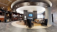 Located on Walnut Street in the heart of Philadelphia, this single-floor space showcases a refined, contemporary interior defined by a striking circular coffered ceiling with integrated lighting at its center. The dramatic ceiling detail creates a natural focal point over the main lounge area, which features a sleek fireplace flanked by a wall-mounted flat-screen display. Comfortable seating arrangements, including leather armchairs and upholstered sofas accented with warm-toned throw pillows, are anchored by a decorative area rug that complements the neutral tiled flooring. The circular design motif flows seamlessly throughout the layout, guiding visitors from the central lounge toward a more intimate dining or bar area to the left, where booth-style seating is illuminated by stylish pendant lighting against a darker, moody backdrop. The overall palette blends charcoal, warm gray, and earthy amber tones, creating an atmosphere that is both polished and inviting. Industrial-influenced architectural elements, such as exposed structural details and dark metal finishes, add depth and character to the space, resulting in an ambiance that balances modern sophistication with approachable comfort.
