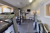 This dedicated fitness room offers a fully outfitted workout environment designed to support a wide range of exercise routines. The space is finished with durable rubber flooring throughout, providing a safe and practical surface for all types of training activity. Neutral gray walls and recessed lighting, complemented by a ceiling fan, create a comfortable and functional atmosphere. Multiple windows fitted with horizontal blinds allow natural light to fill the room while offering privacy, and a wall-mounted clock keeps workouts on schedule. The equipment selection is extensive and well-organized, including a multi-station cable and weight machine for comprehensive strength training, a treadmill, an elliptical trainer, a rowing machine, and a flat weight bench. A wall-mounted television provides entertainment during cardio sessions. A built-in window ledge along one wall serves as a convenient surface for free weights, including a set of dumbbells. The room features an interior pass-through window connecting to an adjacent space, along with two access doors. Thoughtful layout and a variety of equipment make this fitness room a fully functional space for cardiovascular conditioning, strength building, and flexibility training alike.
