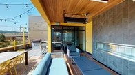 This rooftop terrace is a well-appointed outdoor amenity space designed to balance shelter and open-air enjoyment. The layout is divided into distinct functional zones, each offering a unique experience. A partially covered lounge area anchors the space, featuring a warm wood-paneled ceiling, overhead infrared heaters, and a striking floor-to-ceiling fireplace clad in chevron-patterned charcoal tile. Blue cushioned seating surrounds the fireplace, creating an inviting gathering spot. Bold yellow structural columns and paneling provide a lively visual identity throughout the terrace.

An open-air bar counter crafted from warm wood runs along the perimeter railing, lined with yellow metal barstools and offering unobstructed panoramic views of the surrounding urban skyline. String lights add a festive ambiance across the open sections of the deck. A dedicated outdoor dining area with a table and coordinating yellow chairs sits adjacent to a gas grill station, making the space well-suited for entertaining.

The deck surface throughout is composed of warm-toned hardwood tiles, complementing the natural wood ceiling of the covered zone. Glass doors connect the terrace to an interior space, ensuring seamless indoor-outdoor flow. The combination of functional amenities, durable materials, and thoughtful design makes this rooftop terrace a standout feature.