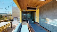 This rooftop terrace is a well-appointed outdoor amenity space designed to balance shelter and open-air enjoyment. The layout is divided into distinct functional zones, each offering a unique experience. A partially covered lounge area anchors the space, featuring a warm wood-paneled ceiling, overhead infrared heaters, and a striking floor-to-ceiling fireplace clad in chevron-patterned charcoal tile. Blue cushioned seating surrounds the fireplace, creating an inviting gathering spot. Bold yellow structural columns and paneling provide a lively visual identity throughout the terrace.

An open-air bar counter crafted from warm wood runs along the perimeter railing, lined with yellow metal barstools and offering unobstructed panoramic views of the surrounding urban skyline. String lights add a festive ambiance across the open sections of the deck. A dedicated outdoor dining area with a table and coordinating yellow chairs sits adjacent to a gas grill station, making the space well-suited for entertaining.

The deck surface throughout is composed of warm-toned hardwood tiles, complementing the natural wood ceiling of the covered zone. Glass doors connect the terrace to an interior space, ensuring seamless indoor-outdoor flow. The combination of functional amenities, durable materials, and thoughtful design makes this rooftop terrace a standout feature.