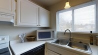This single-floor residence offers a comfortable and practical layout designed for everyday living. The kitchen is a highlight of the space, showcasing classic white cabinetry with gold hardware that provides generous storage both above and below the countertops. Clean white countertops offer ample workspace, while a stainless steel double-basin sink sits beneath a window fitted with horizontal blinds, allowing natural light to filter in while offering a pleasant outdoor view of surrounding trees. A pendant light with a warm bell-shaped shade hangs above, adding a touch of charm to the space. The kitchen is equipped with essential appliances, including a built-in black dishwasher and a countertop microwave, making the space both efficient and convenient. The neutral wall tones complement the white cabinetry, creating a bright and clean aesthetic throughout. Decorative touches such as a woven basket on the counter add a warm, homey feel. The overall ambiance of the space is welcoming and practical, with a timeless design that balances functionality and comfort seamlessly.