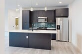 Located at Vintage Commerce in Fort Myers, Florida, this contemporary single-floor unit showcases a thoughtfully designed open-concept layout with consistent light wood-tone flooring throughout. The kitchen serves as the centerpiece of the living area, featuring dark slate cabinetry with gold hardware, a teal subway tile backsplash, stainless steel appliances, and a generous island with a white quartz countertop, undermount sink, and stylish pendant lighting. Recessed lighting complements the clean white ceilings, creating a bright and modern atmosphere. The bedrooms are well-proportioned, finished with the same light flooring, ceiling fans, and large windows fitted with horizontal blinds that allow natural light while maintaining privacy. Several bedrooms offer views of the surrounding landscape, including palm trees. A full bathroom features a floating white vanity with gold hardware, a framed black mirror, matte black fixtures, and a tiled tub-and-shower combination. A private balcony accessible from the main living area offers open-air outdoor space with metal railing and views of the Florida surroundings. The overall design aesthetic is clean, modern, and move-in ready.
