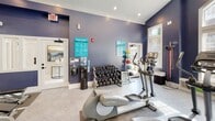 Located in Charlotte, NC, this single-floor fitness center is thoughtfully designed to provide a comprehensive workout environment. The space is defined by its striking deep blue-purple walls, crisp white trim, and vaulted ceilings that lend a sense of openness and volume throughout. Large arched windows and multiple standard windows flood the interior with abundant natural light, offering views of the surrounding greenery and creating an inviting, energizing atmosphere.

The fitness center is organized to accommodate a wide range of exercise disciplines. The cardio zone is well-stocked with multiple treadmills, elliptical trainers, and a stationary cycling bike, with a wall-mounted television nearby for entertainment during workouts. The strength training area features a multi-station cable machine, adjustable benches, and a full rack of dumbbells, catering to users of all fitness levels. A water cooler station and posted informational signage add practical convenience throughout the space.

Full-length wall mirrors visually expand the room and allow users to monitor their form during exercise. The light-toned patterned flooring complements the bold wall color, while recessed lighting and ceiling fans ensure the space remains comfortable and well-lit. The overall design balances functionality with an aesthetically polished environment, making this fitness center an appealing and well-appointed facility for residents.