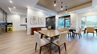Located in Kanata, Ottawa, this contemporary amenity lounge is a thoughtfully designed communal space spread across a single floor. The open-concept layout is anchored by warm light oak hardwood flooring and crisp white walls, creating a bright and welcoming atmosphere throughout.

The space features two well-appointed kitchen areas, each outfitted with sleek dark wood cabinetry, white quartz countertops with marble-effect backsplashes, and stainless steel fixtures. One kitchen includes built-in wall ovens, adding functional versatility for gatherings and events.

Multiple dining tables with mid-century modern wooden chairs and upholstered seats are arranged to accommodate a variety of group sizes, making the space ideal for both casual meetings and larger social events. A lounge area with neutral-toned seating provides a relaxed retreat within the room.

The recreation zone is highlighted by a full-sized pool table positioned beneath a wall-mounted flat-screen TV, offering an entertaining focal point. A bold navy feature wall with geometric detailing adds a dramatic design element near the main entrance. Overhead, a slatted wood ceiling detail with pendant lighting distinguishes the dining zone, adding warmth and visual interest. Large glazed doors allow natural light to filter in and provide views to an outdoor area, further enhancing the open and airy ambiance of this well-curated amenity space.