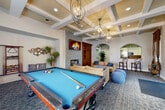 Located at 4656 Quigg Dr in Santa Rosa, California, this single-floor community amenity center offers a thoughtfully designed space for recreation, fitness, and socializing. The centerpiece of the facility is a stylish clubhouse lounge featuring a striking floor-to-ceiling tile fireplace, wall-mounted display, and coffered ceilings adorned with sculptural orb chandeliers. Plush leather sofas and a rustic wood coffee table create a warm, inviting gathering area, while a full-size billiards table adds a recreational focal point to the room. An arched entryway connects the lounge to a well-equipped kitchen, complete with granite countertops, white shaker cabinetry, stainless steel appliances, and a gas range with a stainless hood. A dedicated business center provides a built-in wraparound desk with light wood countertops, hardwood flooring, and natural light from a large window. The fitness center is generously equipped with Cybex cardio machines, strength equipment, a full-length mirror wall, ceiling fans, and abundant natural light from surrounding windows. Multiple hallways connect the various areas throughout the facility. Outdoors, a large pool area with lounge seating and lush landscaping completes this comprehensive amenity offering.