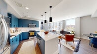 This contemporary single-floor residence showcases a thoughtfully designed open-concept layout that seamlessly connects the kitchen, dining, and living areas. The kitchen is a standout feature, equipped with bold blue cabinetry, white quartz countertops, a white subway tile backsplash, stainless steel appliances, and a central island with pendant lighting — ideal for both cooking and casual dining. Hardwood flooring runs throughout, adding warmth and continuity.

The living area is bathed in natural light courtesy of expansive floor-to-ceiling windows that frame sweeping urban views, creating an airy, elevated atmosphere. The space accommodates comfortable seating and opens naturally to the rest of the home.

Two well-appointed bedrooms provide restful retreats. The primary bedroom features a built-in closet with integrated shelving and drawers, direct access to an en-suite bathroom with a glass-enclosed walk-in shower, and generous natural light from large windows with city views. The second bedroom also benefits from large windows and closet access.

Both bathrooms are finished with light wood vanities, white quartz countertops, and black hardware accents. One includes a tub-shower combination, while the other features a frameless glass shower. Clean, neutral tones throughout create a cohesive and modern aesthetic.