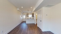 This two-floor residence showcases a clean, contemporary design with thoughtfully selected finishes throughout. The main level opens into a spacious living and kitchen area anchored by rich, dark hardwood-style flooring. The kitchen is a standout feature, outfitted with gray shaker-style cabinetry, white quartz countertops, a black matte sink faucet, stainless steel appliances including a dishwasher and refrigerator, and a central island with pendant lighting that adds both function and style. Recessed lighting complements the natural light flowing in through a front entry window and door. A half bathroom on the main level features hardwood-style flooring and matte black hardware, maintaining a consistent design language. The upper level includes multiple bedrooms finished with plush carpeting, each benefiting from natural light through windows with neighborhood views. One bedroom features a ceiling fan and direct access to an adjoining bathroom. The bathrooms are finished with white vanities, quartz countertops, and matte black hardware accents. Throughout the space, a neutral color palette of warm whites and soft creams creates a bright, welcoming ambiance suited to a variety of living styles.