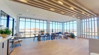 This well-appointed amenity lounge occupies a single floor and is defined by its striking architectural design and abundant natural light. Expansive floor-to-ceiling glass curtain walls wrap around multiple sides of the space, framing sweeping panoramic views of the surrounding cityscape and beyond, creating an airy, open atmosphere throughout.

Warm-toned hardwood flooring runs continuously across the entire space, unifying its several distinct functional zones. A formal dining area features a long wood communal table surrounded by modern black dining chairs, ideal for gatherings or collaborative use. Adjacent to the dining area, a sleek wet bar and cabinetry setup with a beverage refrigerator and herringbone tile backsplash provides convenient service amenities.

A lounge seating area near the glass walls showcases a leather sofa with colorful accent pillows, a round coffee table, and layered area rugs, all bathed in natural light. A more intimate nook features built-in navy blue banquette seating anchored by bold artwork and a decorative shelving unit displaying curated accessories.

The ceiling is a standout architectural feature, adorned with linear wood slat detailing and recessed lighting that adds warmth and visual texture. Vertical wood slat partitions further delineate areas while maintaining an open, cohesive feel. The overall ambiance is modern, sophisticated, and welcoming.