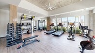 Located at 265 Avenue E SW in Winter Haven, Florida, this fitness center is a thoughtfully designed exercise space that combines modern aesthetics with comprehensive workout functionality. The room features soaring ceilings with an exposed concrete finish, complemented by a large ceiling fan and track lighting that illuminate the space effectively. Warm hardwood-style flooring runs throughout, creating a clean and inviting atmosphere.

Large floor-to-ceiling windows line the walls, flooding the interior with abundant natural light and offering open views of the surrounding greenery and outdoor scenery. The equipment selection is diverse and well-organized, including multiple Spirit treadmills, elliptical machines, a stationary spin bike, and a multi-functional cable machine for strength training. A dedicated TRX suspension training rack is mounted to the wall alongside kettlebells, a medicine ball, and free weights, supporting a full range of functional fitness routines.

A standout feature is the freestanding infrared sauna cabinet, providing a recovery option within the same space. A large flat-screen TV and wall mirrors enhance the workout environment, adding both entertainment and a sense of expanded space. The overall ambiance is modern, bright, and practical, offering a well-rounded fitness experience.