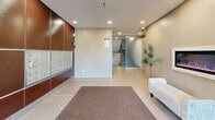 The lobby at 16 Wilsonview Ave in Guelph, Ontario offers a clean, contemporary first impression with a thoughtfully designed layout that balances function and style. The entrance features full-height glass doors framed in sleek black, allowing natural light to filter in and providing a clear view of the surrounding greenery outside. A durable runner mat leads residents from the entry into the main lobby area, finished with large-format light tile flooring that extends throughout the space.

The main lobby is finished with a neutral palette of warm beige and cream tones, complemented by rich dark-paneled accent walls that add depth and visual contrast. Recessed lighting illuminates the space evenly, while a security camera ensures resident safety. A wall-mounted bank of individual mailboxes serves residents conveniently near the entrance, and a cushioned bench provides a comfortable spot for waiting.

Potted ornamental trees add a touch of greenery and warmth to the space. A frosted glass partition wall elegantly separates the entry vestibule from the inner corridor, where large beige tiles continue and lead toward the stairwell. The stairwell is accessed through a glass-paneled enclosure, with a grey door providing additional building access. The overall ambiance is polished, secure, and welcoming.