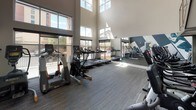 This contemporary fitness center is thoughtfully designed to inspire and support an active lifestyle. The space immediately impresses with its soaring double-height ceilings and an expansive wall of floor-to-ceiling windows that flood the interior with natural light and offer open views of the surrounding neighborhood, creating an inviting and energizing atmosphere.

A striking large-scale athletic mural anchors one wall, depicting dynamic figures in motion — a runner, a boxer, and a cyclist — rendered in a bold graphic style against a deep navy backdrop. This eye-catching feature adds a strong motivational energy to the space.

The fitness floor is generously appointed with a comprehensive selection of cardio equipment, including multiple treadmills, elliptical trainers, stationary bikes, and stair climbers from Life Fitness and other leading brands. A dedicated area near the mural wall houses additional cycling and cross-training machines, alongside colorful medicine balls and exercise accessories stored in an adjacent room.

A sleek open staircase with wood treads and polished steel cable railings adds an architectural element while connecting to an upper-level overlook. A water station is conveniently positioned at the base of the staircase. Full-length mirrors along one wall amplify the sense of space. The durable, textured carpet flooring throughout provides both comfort and function, completing a fitness environment that is as practical as it is visually compelling.