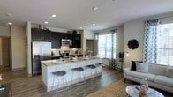 This thoughtfully designed single-floor space showcases a seamless open-concept layout that connects the kitchen and living areas into one cohesive, inviting environment. The kitchen is a standout feature, appointed with rich dark cabinetry that contrasts beautifully against light stone countertops and a classic white subway tile backsplash. Stainless steel appliances, including a full-size refrigerator, microwave, and range, lend a contemporary, professional feel to the space. A generous kitchen island with seating for multiple wire-frame barstools serves as a natural gathering point, ideal for casual dining or entertaining. Recessed lighting throughout the kitchen and living area ensures consistent, warm illumination, complemented by natural light streaming through multiple large windows dressed with patterned curtains. The living area flows naturally from the kitchen, featuring hardwood-style flooring that runs throughout, tying the open space together with a sense of warmth and continuity. Built-in shelving adds functional storage and display opportunities along the walls. The overall ambiance strikes a balance between modern sophistication and comfortable everyday living, making this a well-appointed and highly functional single-level space.