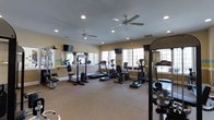 This fitness center offers a comprehensive workout environment designed to support a variety of exercise routines. The space is finished with neutral-toned carpeted flooring and warm beige walls accented by a distinctive yellow and teal horizontal stripe, adding a vibrant touch to the interior.

Natural light filters in through multiple windows along one wall, creating a bright and energizing atmosphere. Recessed ceiling lighting and two ceiling fans ensure consistent illumination and airflow throughout the room, keeping the environment comfortable during workouts.

The facility is equipped with an array of cardio machines, including treadmills, ellipticals, and a stationary cycle, as well as strength training stations featuring cable machines and a multi-function weight rack. Wall-mounted mirrors on one side of the room enhance the sense of space and allow users to monitor their form during exercise.

Entertainment is provided via multiple flat-screen televisions mounted on the walls, ensuring an engaging workout experience. The layout is thoughtfully arranged to provide ample circulation space between equipment, making the fitness center both functional and easy to navigate.
