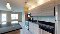This single-floor space showcases a thoughtfully designed open-concept layout that connects the kitchen and living area in a clean, contemporary fashion. The kitchen is outfitted with flat-panel cabinetry in a warm wood-grain finish, providing ample upper and lower storage. Dark stone countertops offer a striking contrast against the lighter cabinetry, and a light blue glass backsplash adds a subtle pop of color. Stainless steel appliances include a range with oven, built-in microwave, dishwasher, and refrigerator, providing a fully equipped cooking environment. A modern faucet with a flexible sprayer is positioned above a deep undermount sink. Track lighting along the ceiling provides focused illumination throughout the kitchen. Light-toned hardwood-style flooring runs throughout, lending warmth and visual continuity between spaces. The adjacent living area benefits from generous natural light streaming through large windows, creating a bright and welcoming atmosphere. A ceiling fan is visible in the living area, adding both function and comfort. Overall, the space presents a clean, modern aesthetic with practical finishes and an airy, open feel.
