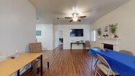This single-floor space offers a thoughtfully arranged layout with warm hardwood-style flooring that flows seamlessly throughout every room, creating a cohesive and inviting atmosphere. The main living area is generously sized, anchored by a classic white-surround fireplace that serves as a striking focal point. A ceiling fan overhead provides both comfort and style, while a wall-mounted flat-screen TV adds a modern touch. Natural light filters in through blinded windows, brightening the open feel of the space. Adjacent to the living area, a dedicated dining space accommodates a dining table and chairs, making it well-suited for gatherings. The galley-style kitchen is efficiently designed with rich cherry wood cabinetry lining both walls, light stone countertops, and white appliances including a range, microwave, refrigerator, and dishwasher — offering ample storage and functionality. A separate room serves as a flexible office space, featuring a long built-in desk surface and natural light from a side window, ideal for productivity. The bathroom includes tile flooring, rich wood cabinetry above the toilet, and a tub-shower combination. Throughout the space, neutral walls and consistent flooring contribute to a clean, versatile aesthetic suitable for a variety of uses.
