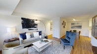 This well-appointed single-floor residence offers a seamless blend of modern design and comfortable living. The living area serves as the centerpiece of the space, featuring warm hardwood flooring that extends throughout, creating a sense of continuity and flow. A sophisticated navy and grey color palette ties the space together, reflected in the plush sofa adorned with coordinating accent pillows and a striking velvet navy armchair with elegant nailhead trim detailing. A bold abstract wall artwork in navy and black tones commands attention above the sofa, lending an artistic focal point to the room. A contemporary arc floor lamp provides ambient lighting, complementing the track lighting visible in the adjoining area. The mirrored coffee table adds a touch of glamour while enhancing the sense of openness. A patterned area rug anchors the seating arrangement, adding texture and depth to the room. The space opens naturally into a dining area, decorated with distinctive wall accents, creating a cohesive entertaining flow. Clean white walls and white interior doors keep the overall aesthetic bright and fresh, while the warm wood flooring grounds the space with an inviting, livable quality.