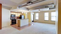 This single-floor urban loft showcases an open-concept layout that seamlessly integrates the kitchen and living areas into one expansive, light-filled space. A defining architectural feature is the exposed metallic ductwork running across the ceiling, lending the space an industrial character that contrasts with the warm, neutral-toned walls and flooring.

The kitchen is well-appointed with light wood cabinetry, a stainless steel refrigerator, and black appliances including a range and microwave. A central island with a breakfast bar serves as both a prep space and a natural divider between the kitchen and the adjacent living area. Pendant lighting hangs above the island, complementing the recessed lighting throughout the kitchen zone. The kitchen area features hardwood-style flooring, while the living space transitions to plush carpeting.

The generous living area benefits from multiple large windows that allow ample natural light to filter in, offering views of the surrounding urban environment. A ceiling fan is centrally positioned, enhancing air circulation throughout. The overall ambiance is modern and functional, blending industrial design elements with comfortable, everyday livability.