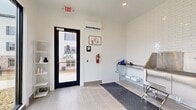 Located at 3415 US-41 in Warner Robins, Georgia, this single-room pet grooming suite — known as Pet Spaw — is a thoughtfully designed, purpose-built space that balances clean aesthetics with professional functionality. The interior is defined by floor-to-ceiling white subway tile on key walls, lending a crisp, hygienic character throughout. Large-format light gray floor tiles provide a durable, easy-to-clean surface underscored by a heavy-duty rubber anti-fatigue mat positioned at the primary work area.

A stainless steel grooming tub with an integrated step, wall-mounted fixtures, and an adjacent stainless steel utility shelf with storage below form the functional core of the space. A separate grooming table with professional-grade equipment completes the workstation setup. A wall-mounted mini-split system ensures climate control year-round.

Natural light pours in through a large black-framed sliding glass door and an expansive picture window, offering outdoor views of the surrounding community. The glass entry door features pet-themed frosted graphics. Compact open shelving along one wall provides additional storage. The overall ambiance is clean, modern, and professionally appointed — well-suited for efficient pet grooming services.