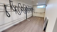 Located at 7911 5th Ave NE in Seattle, WA, this purpose-built bike storage room offers a clean, organized solution for residents' cycling needs. The single-floor space is designed for efficiency and ease of use, featuring a row of wall-mounted vertical bike racks that maximize capacity while keeping the floor area clear and accessible. The racks are constructed from sturdy black metal and are arranged along the full length of the main wall, accommodating multiple bicycles in a space-saving upright position. The room is finished with smooth, wood-toned vinyl plank flooring that is both durable and easy to maintain. Bright recessed lighting ensures excellent visibility throughout the space, while a small window along the upper portion of the wall allows for a degree of natural light and ventilation. The walls are clean and white, contributing to a bright and well-kept atmosphere. A horizontal rail runs along the wall adjacent to the racks, adding a practical element for additional storage or support. The overall layout is straightforward and functional, providing a secure and dedicated environment for bicycle storage that serves the needs of the building's residents efficiently.