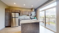 This single-floor space showcases a thoughtfully designed layout with a contemporary aesthetic throughout. The kitchen serves as the focal point, featuring rich dark-toned cabinetry with sleek hardware, granite countertops, and a full suite of stainless steel appliances including a refrigerator, electric range, and built-in microwave. A substantial center island with a matching granite surface and dark base provides additional workspace and casual dining potential. Light hardwood-style flooring runs throughout the kitchen and living areas, complementing the warm, neutral color palette of olive and gray wall tones. A sliding glass door floods the kitchen with natural light and opens directly onto a private outdoor balcony, offering pleasant outdoor views. The bathroom is clean and functional, featuring a full-size soaking tub and shower combo with crisp white surround, chrome fixtures, and coordinating light wood-style flooring. The bedroom presents a well-proportioned, carpeted space with neutral walls, offering a comfortable and versatile environment. Recessed lighting throughout the main living areas adds a polished, modern finish to this well-appointed space.