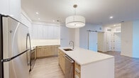 Located at 8005 Gramercy Blvd in Rockville, Maryland, this modern single-floor residence showcases a clean, contemporary design throughout. The open-concept kitchen is a standout feature, outfitted with two-tone cabinetry — glossy white upper cabinets paired with warm natural wood lower cabinets — set against a classic white subway tile backsplash. A large central island with a crisp white quartz countertop provides ample workspace and is complemented by a stylish drum pendant light overhead. Stainless steel appliances, including a refrigerator, range, and over-the-range microwave, complete the well-appointed kitchen. A sliding barn door adds a modern architectural touch off the main living area, which features light hardwood-style flooring found throughout the residence. The living space is bright and airy, with large windows and sliding glass door access to a private balcony overlooking the surrounding community. The bedrooms are generously sized, each offering natural light and closet access. The bathrooms are finished with neutral stone-look tile, featuring a glass-enclosed walk-in shower in one and a full soaking tub with tiled surround in the other, both paired with warm wood vanities and white countertops. The overall ambiance is clean, bright, and move-in ready.