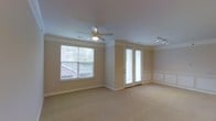 This single-floor space presents a well-appointed living area finished in a warm, neutral color palette that creates a clean and inviting atmosphere. The room is generously sized, offering an open layout with plush wall-to-wall carpeting underfoot. Crown molding runs along the ceiling perimeter, lending a refined, classic character to the space. A ceiling fan with an integrated light fixture is centrally mounted, providing both air circulation and ambient lighting. Track lighting is also installed along one side of the ceiling, allowing for directional illumination. Natural light enters through a large window fitted with horizontal blinds, while a set of French doors with matching blinds provides additional brightness and potential outdoor access. The walls feature partial wainscoting detail on one side, adding a traditional architectural touch. Neutral beige walls and white trim work together to maintain a cohesive and timeless aesthetic throughout. The overall ambiance is calm and understated, with thoughtful finishing details that elevate the everyday functionality of the space.