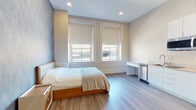 This single-floor studio unit at 9 Peck St in Providence, Rhode Island, presents a thoughtfully designed living space that balances modern aesthetics with practical functionality. The main living area serves as both a sleeping and living space, anchored by a wood-framed bed set against a striking geometric patterned accent wall in cool blue and grey tones, adding a distinctive visual focal point to the room. Light wood-look flooring runs throughout, contributing to a warm, cohesive feel across the unit.

Two large windows fitted with neutral roller shades allow natural light to fill the space while offering privacy, and recessed ceiling lighting supplements the brightness. A built-in desk and a compact stainless steel appliance unit provide convenient workspace and storage directly within the main room.

The kitchenette is efficiently arranged with white cabinetry, a sleek countertop, a stainless steel sink, dishwasher, refrigerator, and microwave — all set against light-toned walls with notably tall ceilings that enhance the sense of openness.

The bathroom features floor-to-ceiling large-format tile in a neutral palette, a glass-enclosed shower/tub combination, and a clean vanity with ample counter space, offering a polished and functional finish.
