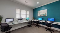 Located at 327 Guinevere Ln in Wilmington, NC, this single-floor space is thoughtfully designed to offer practical amenities in a polished, welcoming environment. The business center serves as the focal point of the space, featuring an L-shaped continuous work surface that accommodates multiple computer workstations along with a multifunction printer. The room is finished with plush carpeting, crown molding, and a bold teal accent wall adorned with an abstract painting, creating a vibrant yet professional atmosphere. White horizontal blinds on the windows allow for natural light control, keeping the workspace comfortable and functional. A reception or concierge nook is also present, outfitted with a built-in desk, a display of informational brochures, and beach-themed wall art, lending a coastal character consistent with the Wilmington locale. Adjacent to the main workspace, a separate room with warm hardwood flooring houses a stand-up tanning bed, set against the same signature teal accent wall. The overall design palette of soft gray walls, white trim, and teal accents creates a cohesive and contemporary look throughout, making this space both efficient and visually appealing.