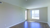 This single-floor space showcases a clean, contemporary design centered around an open living area. The room features rich, dark hardwood-style flooring that contrasts elegantly with the light, neutral-toned walls, creating a warm yet modern aesthetic. Smooth white ceilings complement the minimalist interior, while white baseboards provide a crisp finishing detail throughout.

Natural light is a defining feature of this space, with a large sliding window and a glass-paneled door flooding the interior with abundant daylight. The windows frame an expansive urban skyline view, bringing the outside environment into the living area and enhancing the sense of openness. HVAC vents are discreetly positioned along the upper walls, ensuring functional climate control without disrupting the clean lines of the space.

The layout is straightforward and versatile, offering an open floor plan that can accommodate a variety of furniture arrangements. The combination of plentiful natural light, quality flooring, and urban views makes this a functional and visually appealing space suitable for comfortable everyday living.