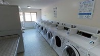 This communal laundry facility is a practical, well-organized space designed to accommodate high-volume laundry needs. The room is laid out with two parallel rows of modern, commercial-grade front-loading washing machines and dryers, all finished in white and equipped with digital control panels for ease of use. The machines are arranged in a clean, efficient configuration that maximizes capacity while maintaining clear pathways for users. The flooring consists of a durable, small-tile mosaic surface well-suited to the demands of a high-traffic environment. Walls are finished in a neutral, light tone that contributes to the room's bright and clean atmosphere. A window on one wall allows for natural light to enter the space, complementing the overhead lighting. Instructional and informational signage is posted on the walls, providing guidance on machine operation. The overall design prioritizes functionality and efficiency, offering residents a straightforward and convenient laundry experience in a tidy, maintained environment.