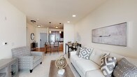 This single-floor residence is designed with an open-concept layout that seamlessly connects the living, dining, and kitchen areas, creating a cohesive and functional living environment. The living room is bright and welcoming, finished with plush carpet and neutral-toned walls. Large sliding glass doors flood the space with natural light and provide access to an outdoor balcony, enhancing the sense of openness. Recessed lighting and pendant fixtures add warmth throughout the shared living spaces.

The kitchen is well-equipped with rich, dark wood cabinetry extending to the ceiling for ample storage, stainless steel appliances including a French door refrigerator, built-in microwave, and electric range, all set atop hardwood-style flooring. A raised breakfast bar with seating connects the kitchen to the dining area, making it ideal for both casual meals and entertaining.

The bedroom is a calm retreat, featuring plush carpeting, generous natural light from a large window with sheer draping curtains, and ample space for a full furniture arrangement including a dresser and desk area. The bathroom offers tile flooring, a white vanity with dark cabinetry, and a closet with paneled gray doors, rounding out the functional and tastefully designed interior.