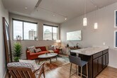 This contemporary urban residence is thoughtfully designed across a single floor, offering a seamless blend of style and functionality. The open-concept layout connects the living area and kitchen, creating a bright and airy atmosphere enhanced by track lighting overhead and warm hardwood-style flooring that runs throughout the space.

The living area is anchored by large windows that frame striking city skyline views, allowing abundant natural light to fill the room. Pendant lighting adds a refined touch to the space, complementing the modern aesthetic.

The kitchen is well-appointed with rich dark wood cabinetry extending to the upper walls, a suite of stainless steel appliances including a full-size refrigerator, gas range, and microwave, and a sleek white quartz peninsula with bar seating — ideal for casual dining or entertaining. Architectural framed prints adorn the kitchen walls, adding a distinctive urban character.

The bedroom is a calm and comfortable retreat, featuring neutral tones, warm ambient lighting, and a wall-mounted mirror that enhances the sense of space. The room accommodates a full-size bed with ease and maintains a clean, uncluttered design.

The bathroom includes dark wood cabinetry, a white quartz vanity countertop, and a full shower enclosure with sliding glass doors, offering both style and practicality. The consistent use of hardwood-style flooring ties the entire residence together cohesively.