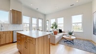 This contemporary single-floor residence showcases a thoughtfully designed open-concept layout that seamlessly connects the kitchen, dining, and living areas. The kitchen is outfitted with warm wood-toned cabinetry, a white quartz-topped island, stainless steel appliances, and a tile backsplash — all set against light hardwood flooring that extends throughout the home. A dining nook adjacent to the kitchen provides a casual space for meals, benefiting from the same natural light that fills the open living area. The living room is bright and airy, featuring multiple large windows that frame outdoor views and flood the space with sunlight. The primary bedroom is a calm retreat with a well-proportioned window, clean white walls, and access to a dual-sink bathroom finished with wood cabinetry and white quartz countertops. A spacious walk-in closet with wrap-around hanging rods offers generous storage. The second bathroom mirrors the same modern aesthetic, complete with a single-sink vanity, a glass-enclosed shower, and light wood finishes. Throughout the residence, the consistent use of warm wood tones, white surfaces, and light flooring creates a cohesive, contemporary ambiance that feels both inviting and functional.