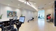 This contemporary fitness center in Dallas is designed to offer a clean, motivating environment for a full range of workouts. The space is spread across a single floor and features a bright, open layout with light hardwood-style flooring and crisp white walls that create an airy, welcoming atmosphere.

The main workout area is equipped with Expresso interactive cycling bikes, each outfitted with large touchscreen displays for an engaging, tech-forward cardio experience. A rowing machine is centrally positioned, providing a versatile full-body workout option. Large ceiling fans with modern fixtures are mounted above, ensuring comfortable airflow throughout the space.

A connected room, visible through floor-to-ceiling glass partitions, houses additional cardio equipment including treadmills, allowing natural light to filter through and giving the overall space a sense of openness and continuity. Recessed lighting throughout ensures consistent illumination at any hour.

Decorative touches such as a colorful abstract wall painting and an accent plant add visual warmth without distracting from the functional purpose of the space. The thoughtful layout ensures ample room between equipment, promoting a safe and comfortable workout environment. Overall, this fitness center balances modern design with practical amenities to deliver a high-quality exercise experience.