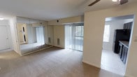 This single-floor residential space offers a clean, functional layout designed for comfortable everyday living. The main room is finished with wall-to-wall neutral-toned carpeting, lending a warm and quiet atmosphere underfoot. A ceiling fan is positioned overhead, providing efficient air circulation throughout the space. One wall features full-length mirrored closet doors, which visually expand the room while offering generous built-in storage. Large sliding glass doors dressed with vertical blinds allow for plentiful natural light and provide a direct connection to the outdoors, with views of the surrounding exterior. An additional window further brightens the space, reinforcing an open and airy feel. The walls are finished in a neutral beige tone, creating a versatile backdrop well-suited to a variety of interior styles. A tiled area is partially visible at the edge of the room, suggesting a transition to an adjacent space such as a bathroom or entryway. Wall vents indicate a central HVAC system for year-round climate control. Overall, the space presents a straightforward, well-maintained interior that balances practicality with comfort.