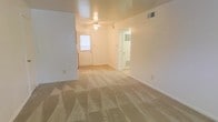 This single-floor residential space presents a clean, functional layout with a warm and neutral aesthetic throughout. The main living area is generously proportioned, featuring wall-to-wall carpet flooring in a light beige tone that adds a sense of comfort underfoot. Smooth, light-colored walls and a flat ceiling create a simple, understated interior palette.

A ceiling fan is centrally positioned in the living area, providing both ventilation and a subtle design element. Natural light enters through a paneled front door with a window, brightening the space and giving it an open, airy feel. A ceiling-mounted light fixture supplements the natural light, ensuring the room remains well-lit throughout the day.

Visible HVAC vents integrated into the walls and ceiling indicate a climate-controlled environment. A hallway or secondary doorway leads further into the space, suggesting additional rooms beyond the main living area. White baseboards and clean wall transitions add a crisp, finished appearance throughout.

Overall, the space offers a straightforward, move-in-ready environment with a neutral canvas well-suited for a variety of interior styles and furnishing arrangements. The open layout and consistent finishes give the space a cohesive and tidy character.