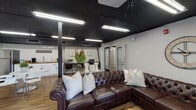 Located at 500 E Hargett St in the heart of Raleigh, NC, this single-floor commercial space offers a thoughtfully designed environment ideal for gatherings, events, or collaborative use. The interior blends industrial and modern aesthetics, highlighted by a striking matte black ceiling with exposed infrastructure and bright linear lighting that illuminates the open floor plan below.

The main lounge area is anchored by a large tufted leather sectional sofa, a cowhide area rug, and a live-edge coffee table, creating a warm and inviting atmosphere. A ceiling-mounted projector paired with a large built-in projection screen makes the space well-suited for presentations or screenings. Windows along one wall allow natural light to flow in, offering views of the surrounding neighborhood.

A dedicated coffee bar area features white shaker cabinets, a butcher block countertop, subway tile backsplash, and a full-size refrigerator — providing functional amenities for hosting. A small dining table with modern white chairs sits adjacent to this area, adding versatility to the layout.

The space also includes a private glass-door room and additional flex areas, connected by wood-look plank flooring that runs throughout. The overall ambiance is polished yet relaxed, making it a highly functional and attractive venue in downtown Raleigh.