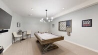 Located in Oak Creek, Wisconsin, this dedicated game room offers a well-appointed recreational space designed for entertainment and leisure. The room features light wood-look flooring and crisp white walls that create a clean, modern aesthetic throughout.

At the center of the space stands a substantial rustic-style pool table with a light felt surface and X-base pedestal design, illuminated overhead by a striking modern chandelier with globe bulbs on a branching black frame. Recessed lighting further brightens the room, adding to its inviting atmosphere.

A large wall-mounted flat-screen television paired with a soundbar anchors one wall, complemented by a dedicated cue rack stocked with billiard cues. Decorative wall-mounted board games, including a large Scrabble board and framed checkerboard art pieces, add a playful, thematic character to the space. A touchscreen gaming table provides an additional interactive entertainment option.

The room is accessed through a sleek black-framed glass door and includes a standard interior door as well, offering convenient access. Seating is available around the perimeter of the room for spectators and participants alike. Overall, the space balances a contemporary design sensibility with a fun, functional layout ideal for communal recreation.