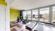 This modern fitness center offers a thoughtfully designed workout space across a single floor. The interior is immediately distinguished by its bold color scheme, featuring vivid chartreuse-yellow accent walls contrasted against deep charcoal panels, creating an energetic and motivating atmosphere. The flooring throughout is a striking chevron-patterned hardwood in a mix of warm and cool tones, adding a dynamic visual element to the space.

Large floor-to-ceiling windows line one wall, flooding the room with abundant natural light while providing open views of the surrounding outdoor landscape, including waterfront scenery and greenery. Recessed ceiling lighting further enhances the brightness of the space.

The fitness center is divided into functional zones. The cardio area is equipped with treadmills, an elliptical trainer, and a recumbent bike. A dedicated strength-training section features a cable machine, a weight bench, and a full dumbbell rack, all complemented by full-length mirrors that expand the sense of space. An entry area with chartreuse walls provides access to an HVAC utility room and includes a water fountain, a waste bin, and a glass-paneled exit door. The overall layout is open and efficiently organized, making this fitness center a practical and visually appealing amenity.