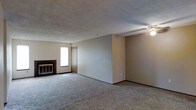 This single-floor residence offers a straightforward and functional layout designed for comfortable everyday living. The main living area is spacious and open, featuring wall-to-wall carpet flooring that adds warmth underfoot. A brick fireplace serves as a focal point along one wall, providing both a practical heating element and a classic architectural detail that anchors the room. Neutral-toned walls keep the space feeling bright and versatile, ready to accommodate a variety of furnishing styles. A ceiling-mounted light fixture provides overhead illumination, complementing the natural light that filters in through a window near the fireplace. The textured ceiling adds a subtle dimensional quality to the interior. A doorway connects the living area to adjacent spaces, suggesting a practical and connected floor plan. The overall ambiance is calm and understated, offering a blank canvas that lends itself well to personalization. The open feel of the living space makes it well-suited for both relaxed daily use and casual entertaining.