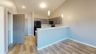 Located at 2300 N Valley View Rd in Sioux Falls, SD, this two-bedroom, one-bathroom unit offers a well-designed layout across a single floor. The open-concept kitchen is a standout feature, boasting vaulted ceilings, gray shaker-style cabinetry, light quartz countertops, and a full suite of black Whirlpool appliances including a refrigerator, range, microwave, and dishwasher. Pendant lighting illuminates the kitchen peninsula, which provides additional prep and seating space. Wood-look plank flooring runs throughout the main living and kitchen areas, adding warmth and continuity. A ceiling fan and a sliding glass door in the living area lead to a private balcony, filling the space with natural light and offering outdoor views. The two bedrooms are carpeted and finished in neutral tones, with windows that bring in additional natural light. The primary bedroom is generously sized and provides convenient access to the full bathroom. The bathroom features a gray vanity with ample storage, a tub/shower combo, and wood-look tile flooring. A dedicated utility room with a water heater rounds out the functional layout. The overall palette of soft grays and neutral tones gives the space a clean, modern aesthetic throughout.
